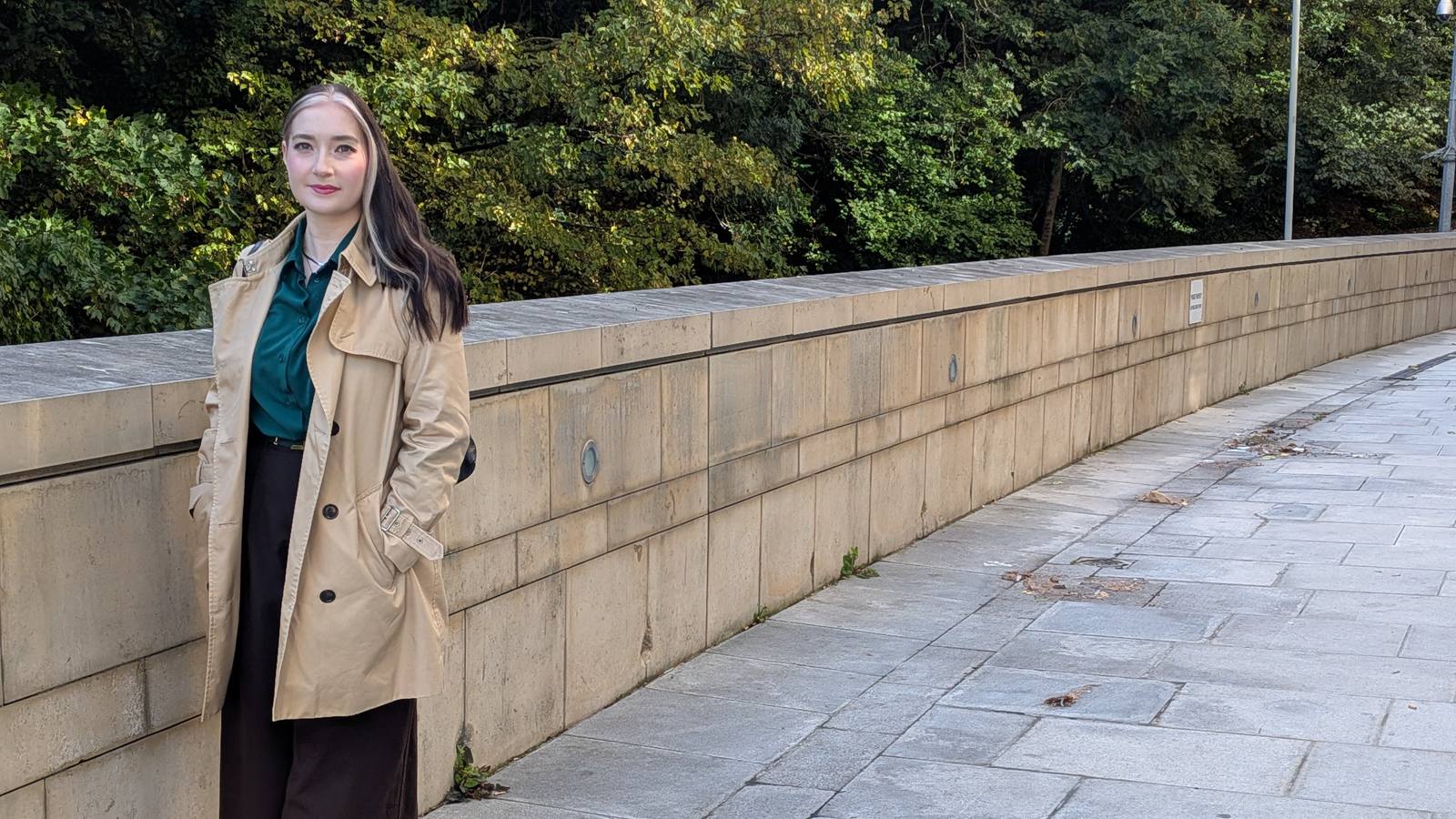 Mirella Fasoula leaning against a stone wall on a quiet, tree-lined path
